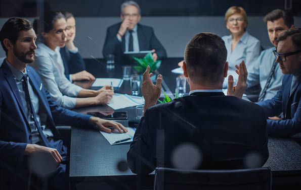 Late at Night In the Corporate Office Meeting Room: At Conference Table Executive Director Talks to a Board of Directors, Investors and Business Associates  Over the Shoulder Shot 